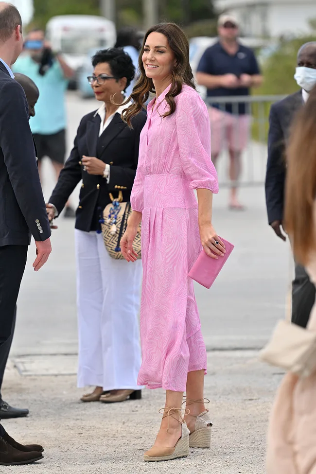Woman in a pink dress and nude heels, holding a pink clutch, outdoors with several people around.