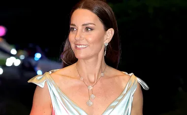 A woman in an elegant gown with statement jewelry, smiling against a dark blurred background.