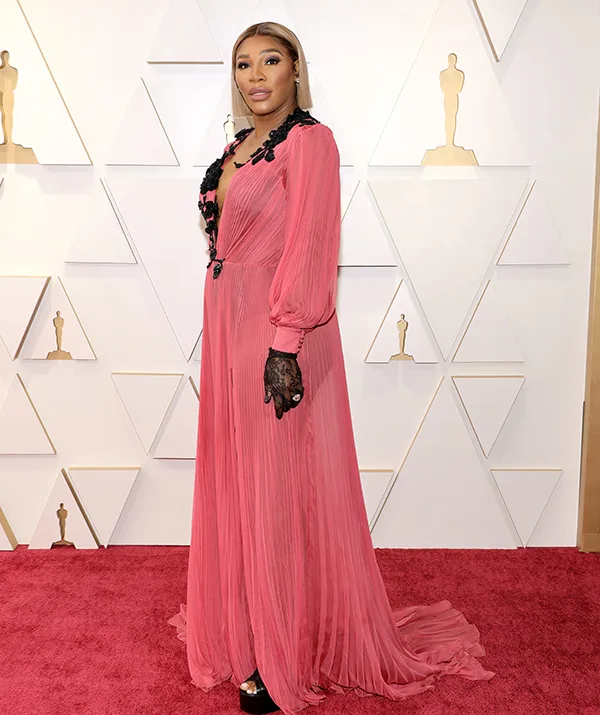 Woman in a pink gown with black lace on a red carpet at a formal event, standing against a geometric backdrop.