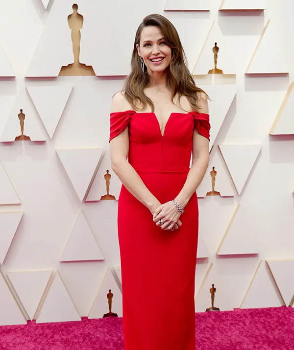 Individual in a red off-shoulder gown on the red carpet at an award event, smiling.