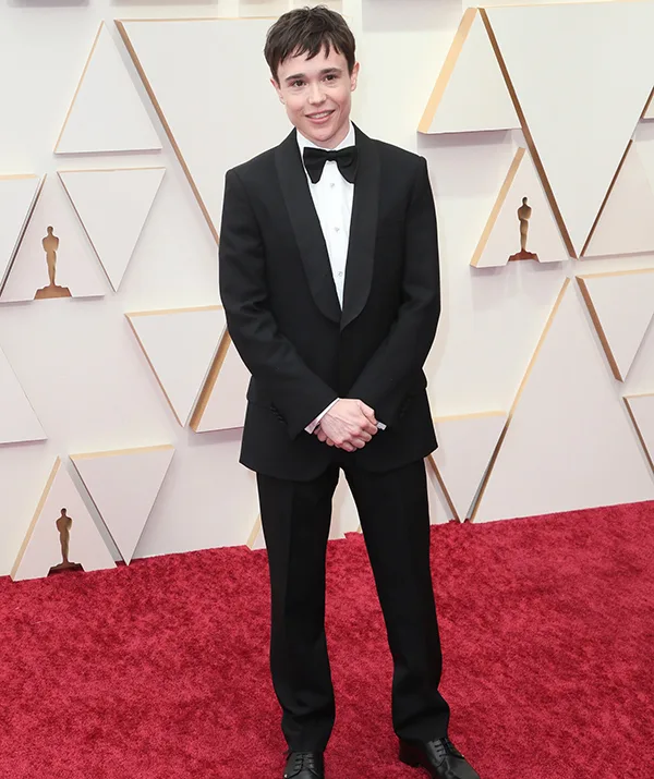 A person in a black tuxedo stands on a red carpet, in front of a geometric-patterned backdrop.
