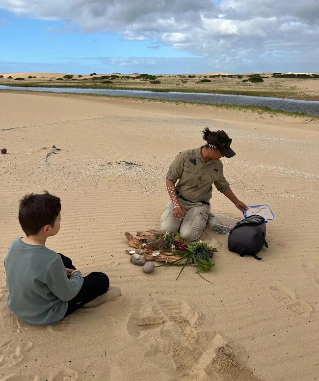 Person demonstrating a cultural activity on a sandy beach, with a child observing.