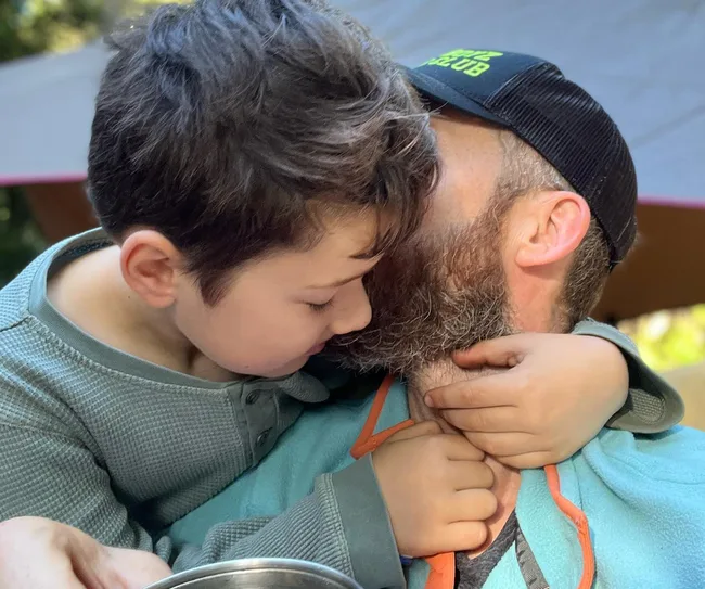 Father and son hugging closely outdoors with tent in background.