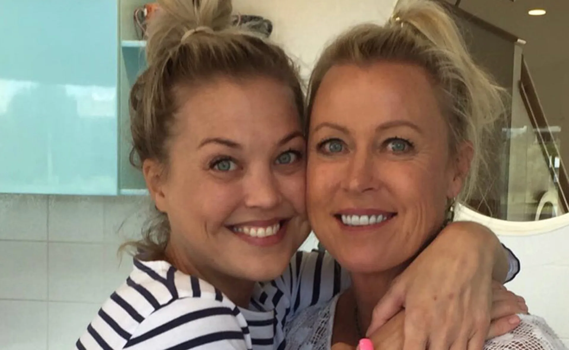 Two women smiling, embracing each other with their hair styled in top knots, standing in a modern kitchen.