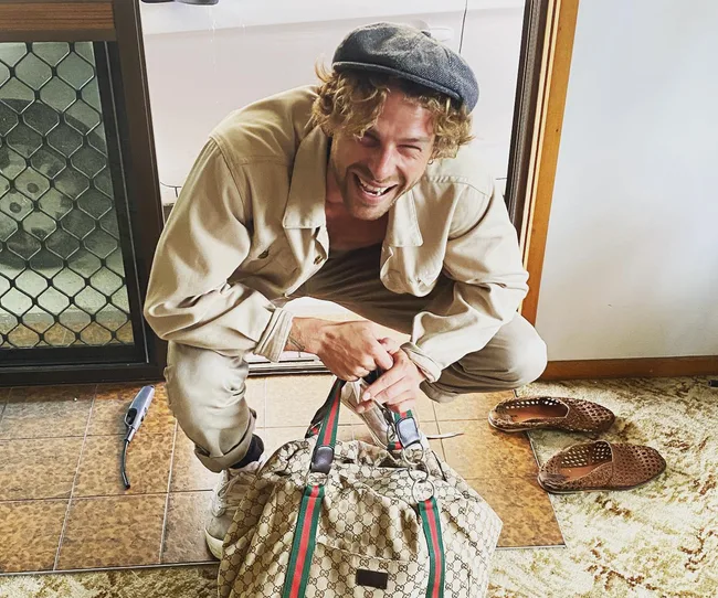 Man smiling, squatting with a Gucci bag, wearing a cap, tan outfit, shoes beside him, in front of a door.