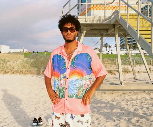 Man in colorful shirt and sunglasses stands on a beach near a lifeguard tower, with sand and grass in the background.
