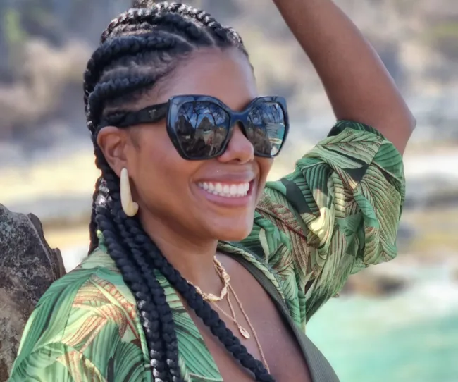 Woman smiling in sunglasses and braids, wearing a green leaf-pattern shirt, outdoors on a sunny day.