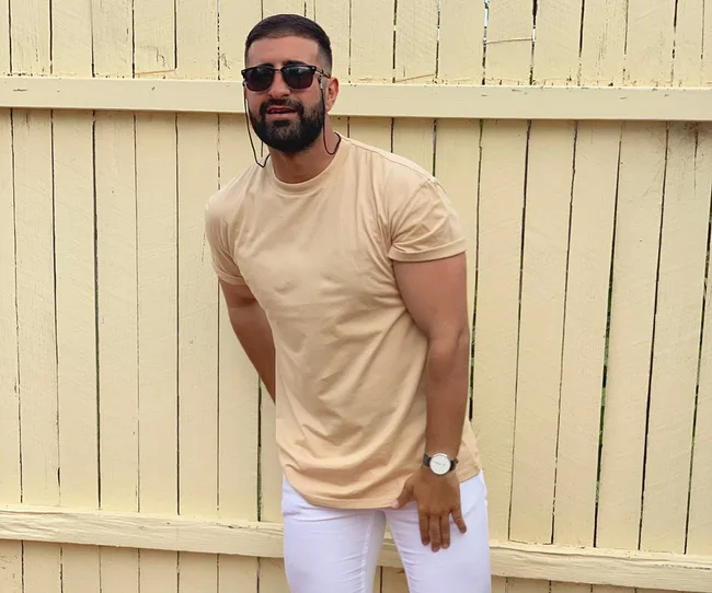 Man wearing sunglasses, beige shirt, and white pants standing in front of a wooden fence.