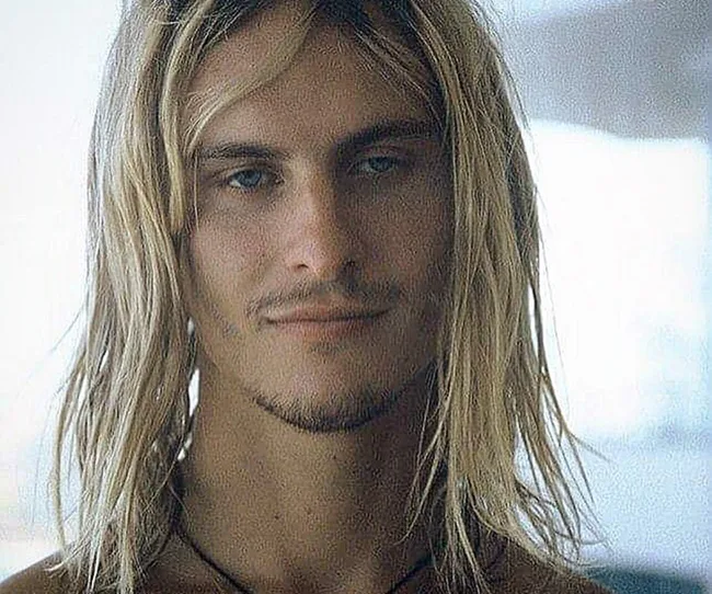 Long-haired person with a slight smile, wearing a necklace, looking directly at the camera in a softly lit setting.