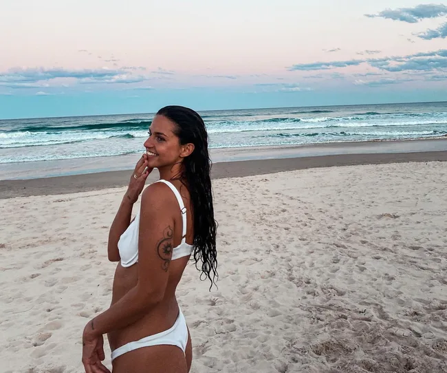 A woman in a white bikini stands on a sandy beach, smiling, with the ocean and sunset in the background.