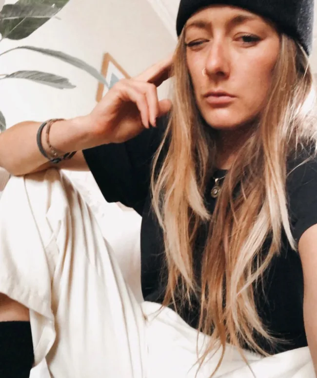 A person with long hair and a nose ring winking, wearing a black beanie and shirt, sitting indoors.