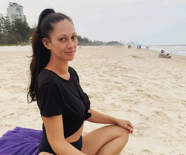 Person sitting on a sandy beach with a towel, wearing a black outfit, with the sea and buildings in the background.