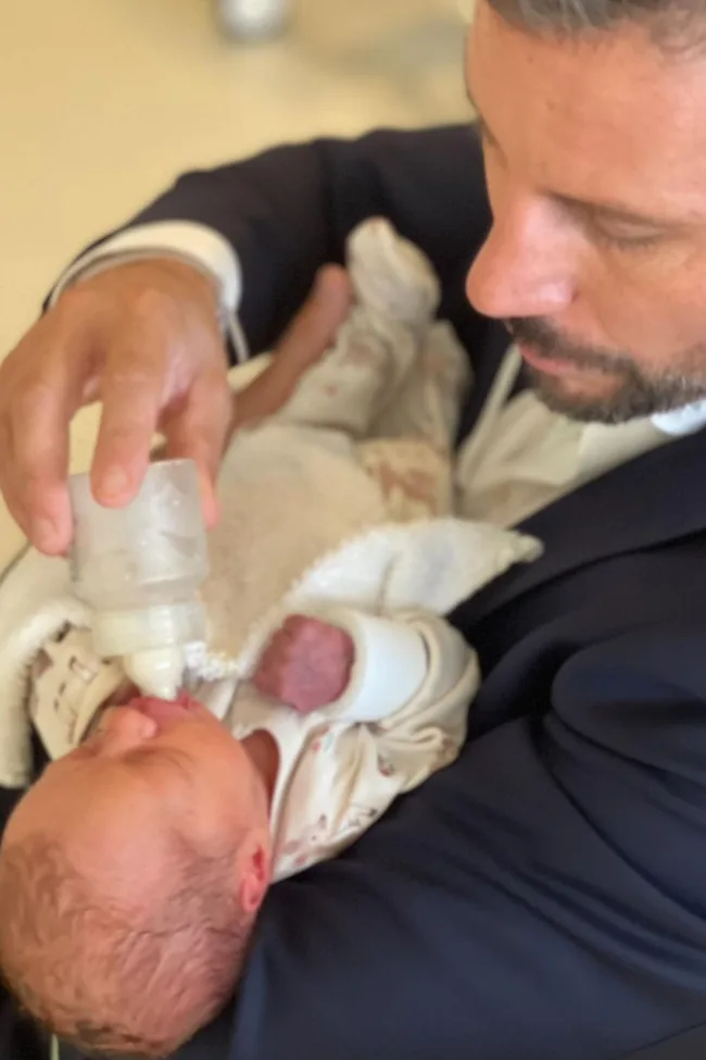 A man in a suit bottle-feeding an infant wrapped in a blanket.