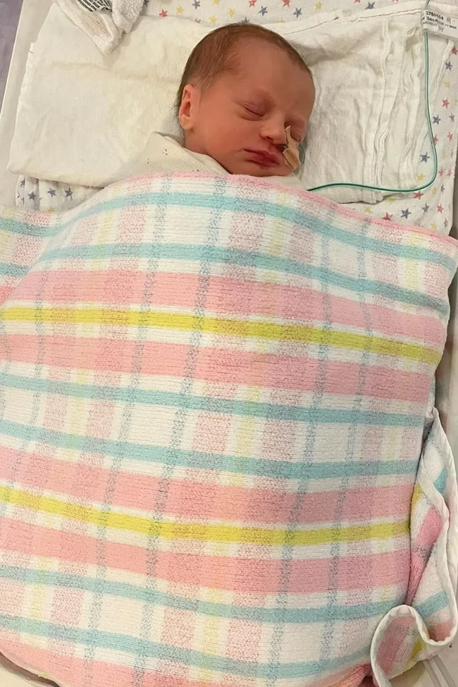 Newborn baby sleeping with a feeding tube, wrapped in a pastel checkered blanket.