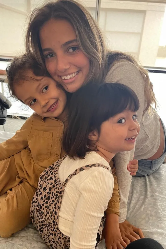 A woman smiling and hugging two young children, all sitting closely together indoors.