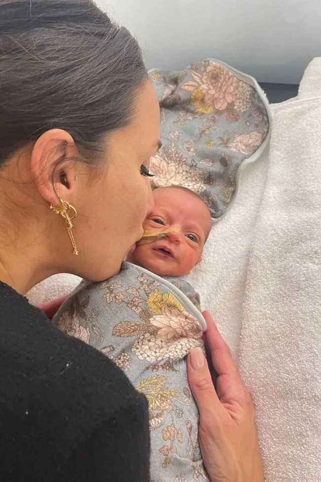 Woman kissing a swaddled baby with a floral blanket, who is wearing a nasal tube.