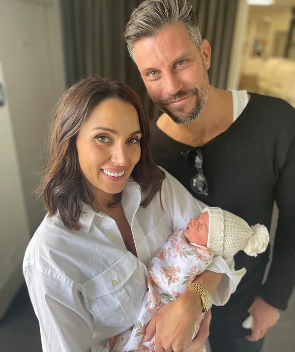 A smiling couple holds a newborn baby wrapped in a floral blanket with a white knitted hat.