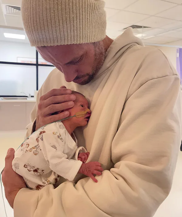 Man in beige hoodie and beanie cradles a baby wearing oxygen tube in hospital setting.
