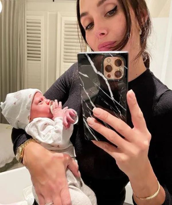 Selfie of a woman in black holding a newborn baby in a white outfit and hat, mirror reflection.