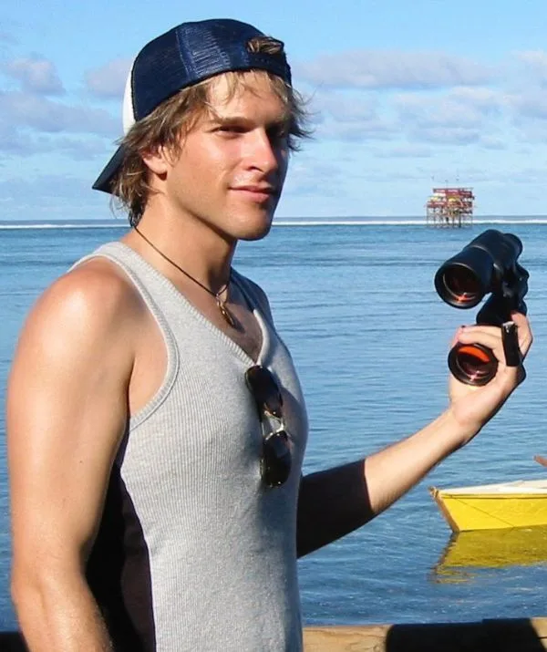 A person in a cap holds binoculars by the water, with a distant platform and boat visible in the background.