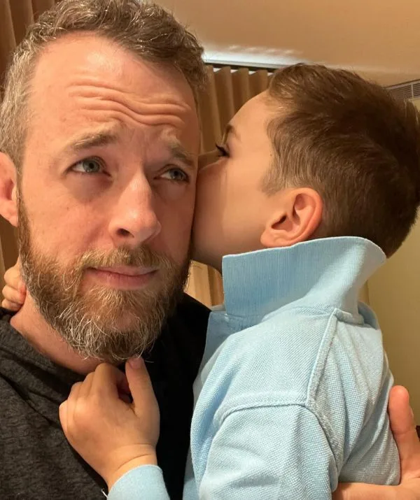 Man with a beard looking thoughtful while a young child whispers in his ear, both indoors.