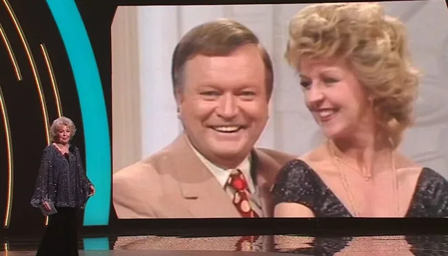 Senior woman on stage with large screen showing a smiling man and woman from an old TV show.