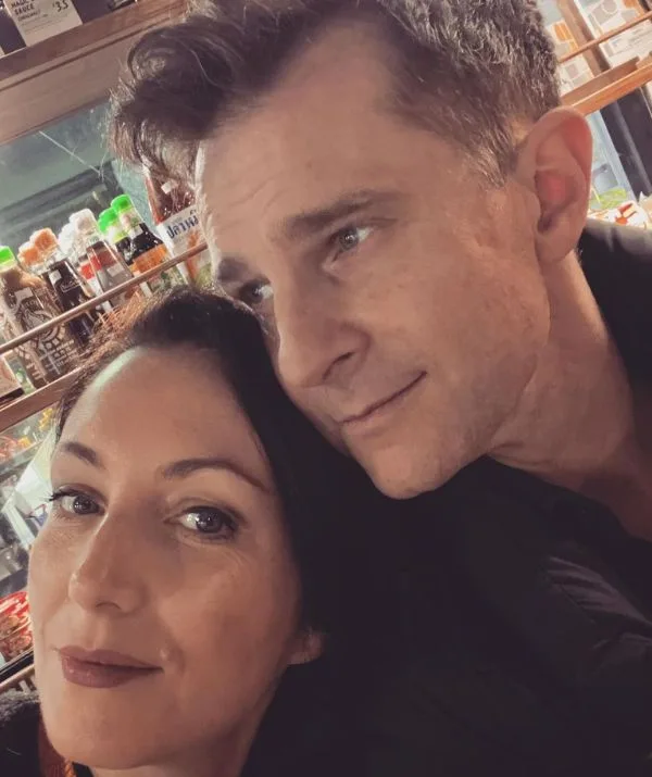 Man and woman taking a selfie indoors, standing close together with shelves of bottles in the background.