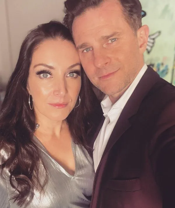 A woman in a silver top and a man in a suit take a selfie together. Both look at the camera with neutral expressions.