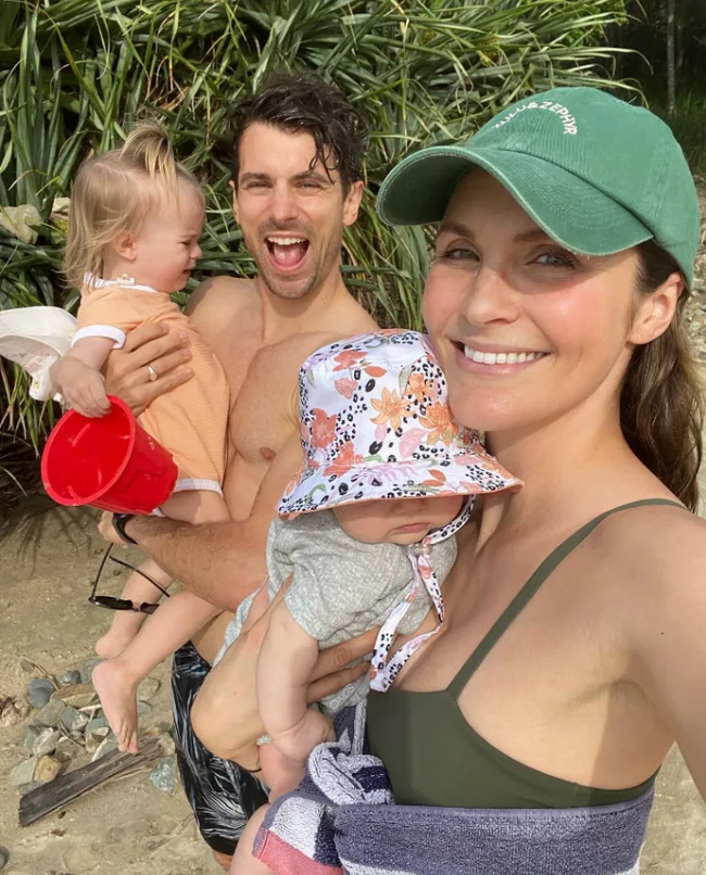 Family enjoying a beach day; woman in green cap, man holding a child with a red bucket, another child in floral hat.