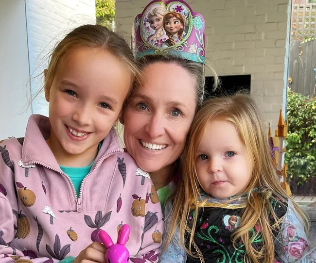 A smiling woman with children wearing colorful outfits, one child in a "Frozen" themed dress, and a "Frozen" crown on the woman.