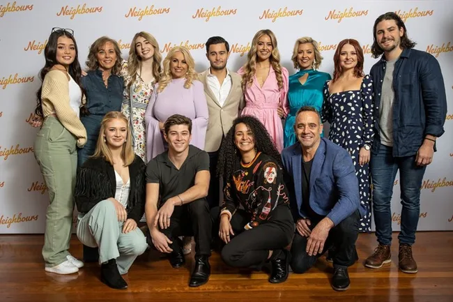 Group photo of "Neighbours" cast members standing in front of a backdrop with the show's logo.