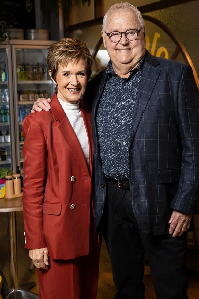 Two people smiling, the woman in a red suit and the man in a blue blazer, standing in a warmly-lit cafe setting.