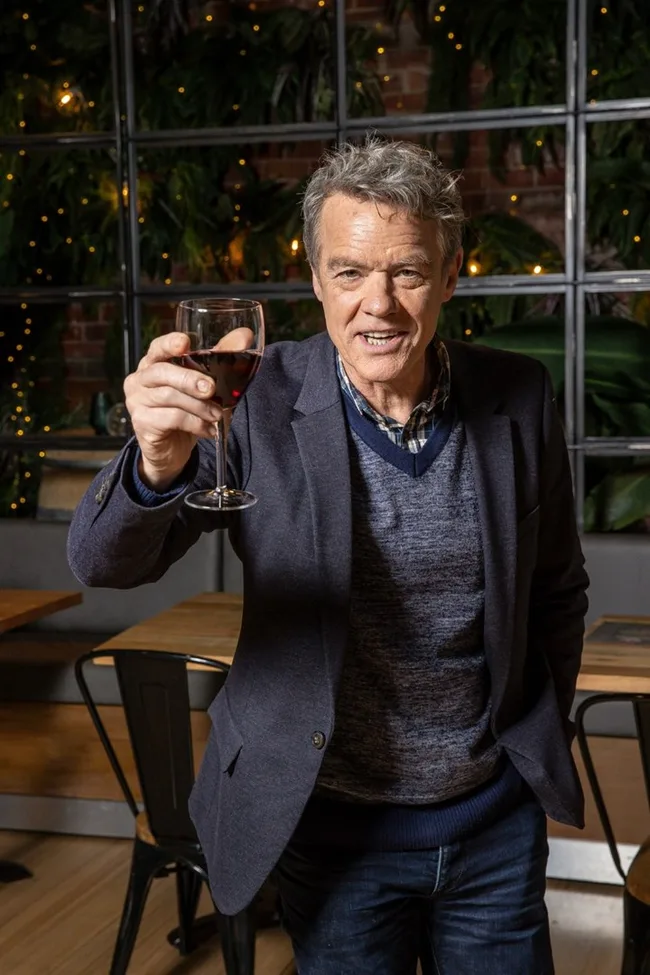 Man in a blue blazer holds a glass of red wine and smiles in a warmly lit room with plants.