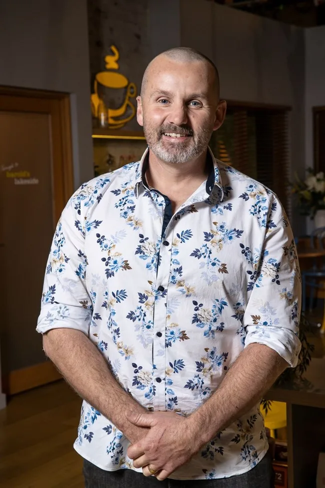 A person wearing a floral shirt, smiling indoors with decorative elements in the background.