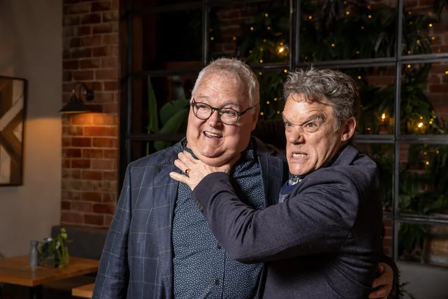 Two men in suits, one playfully holding the other in a mock chokehold, both smiling in a warmly lit room.