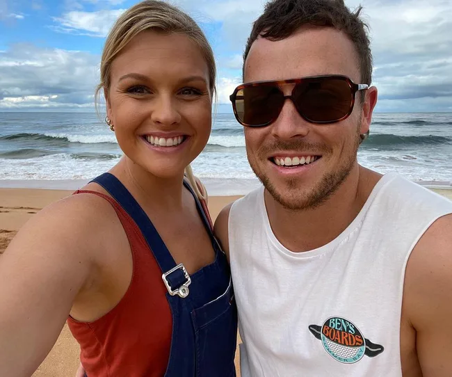 A smiling man and woman take a selfie on a beach with waves in the background, under a partly cloudy sky.