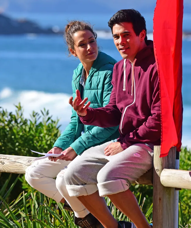 A man and a woman sit on a wooden fence near the ocean, talking, beside a red flag.