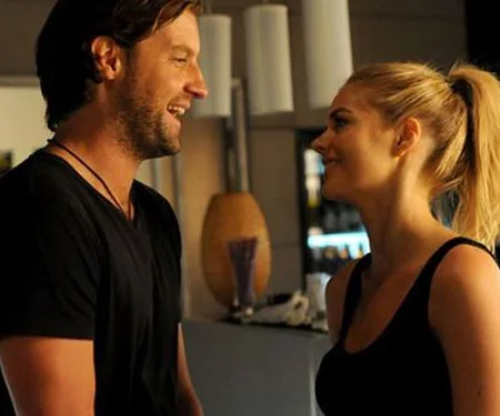 A man and woman smiling at each other in a dimly lit room, suggesting a casual and friendly interaction.