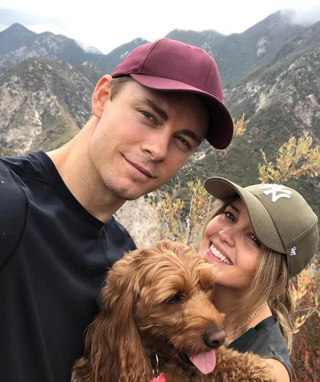 A couple wearing baseball caps smiles with a brown dog in front of mountain scenery.