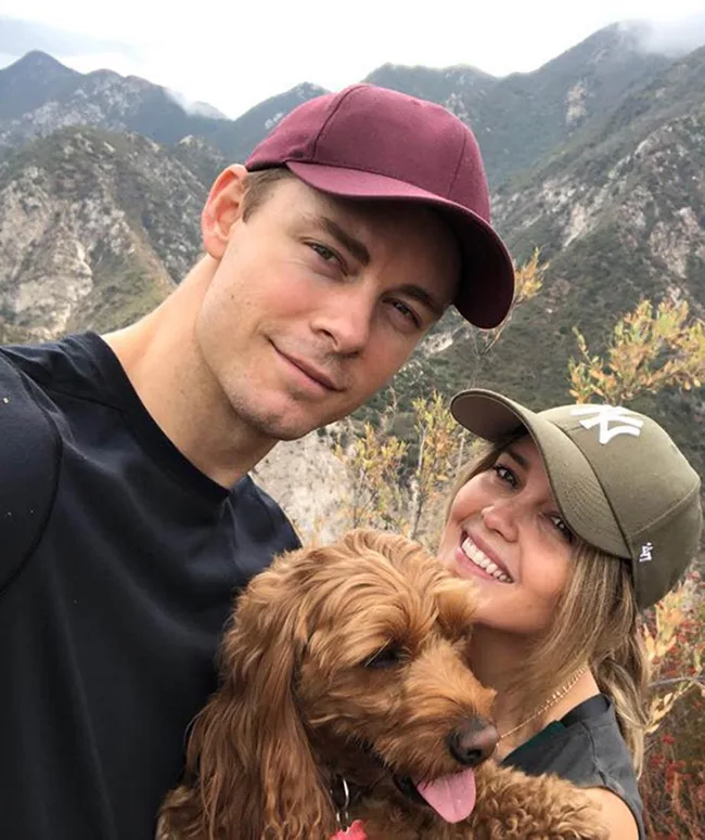 A couple wearing baseball caps smiles with a brown dog in front of mountain scenery.