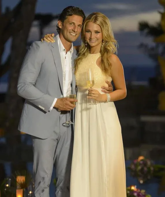A couple in formal attire holding champagne glasses, posing outdoors at sunset.