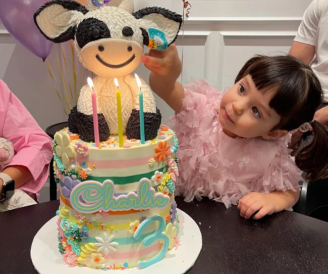 A child in a pink dress admires a colorful birthday cake with a cow topper, candles, and "Charlie 3" on it.
