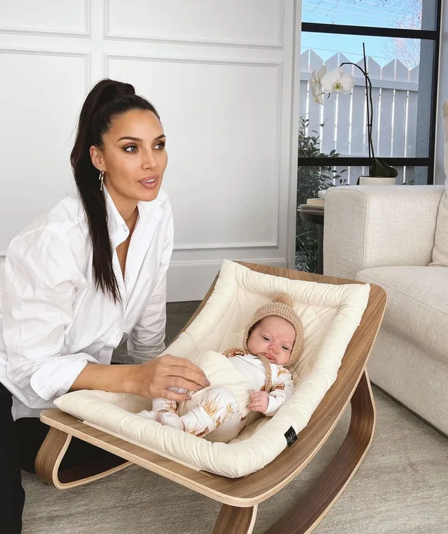 Woman in a white shirt kneeling beside a baby in a wooden rocker chair indoors.