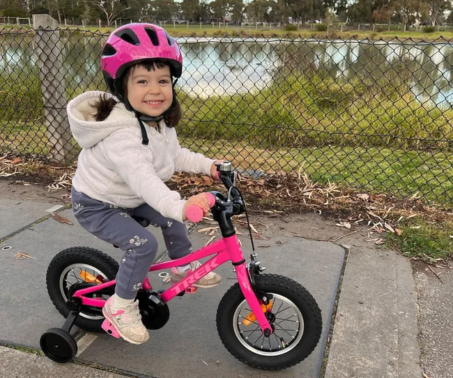 Young child smiles while riding a pink bike with training wheels, wearing a pink helmet, near a chain-link fence by a lake.