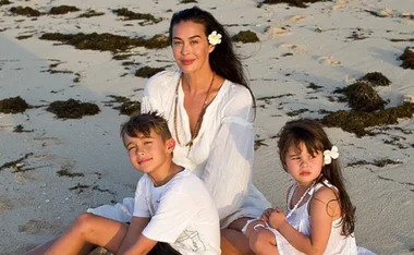 A woman and two children sit on a sandy beach, wearing white outfits with flowers in their hair.