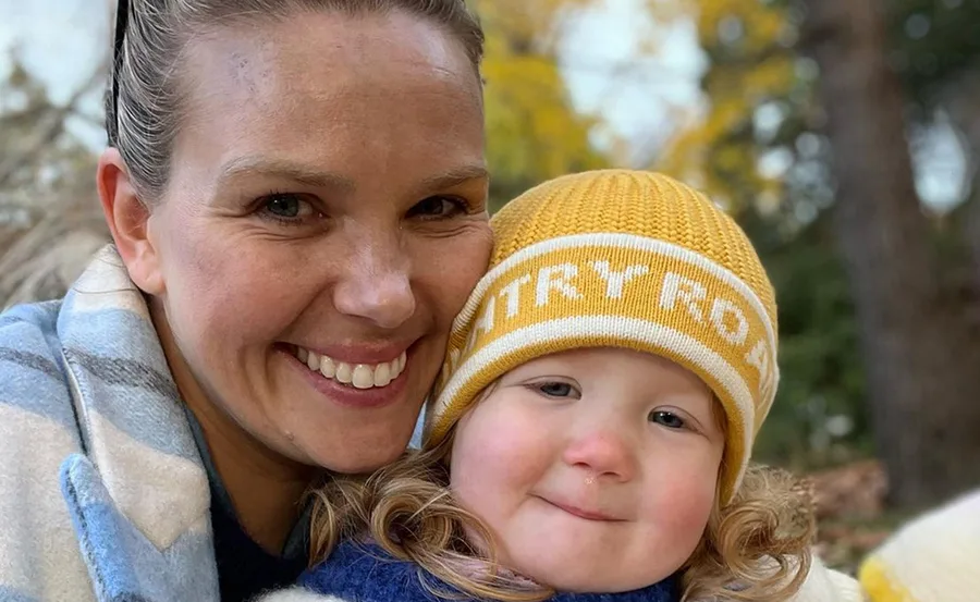 Mother smiling with child in a yellow beanie outdoors, autumn foliage in the background.