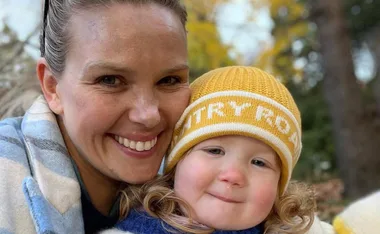 Mother smiling with child in a yellow beanie outdoors, autumn foliage in the background.