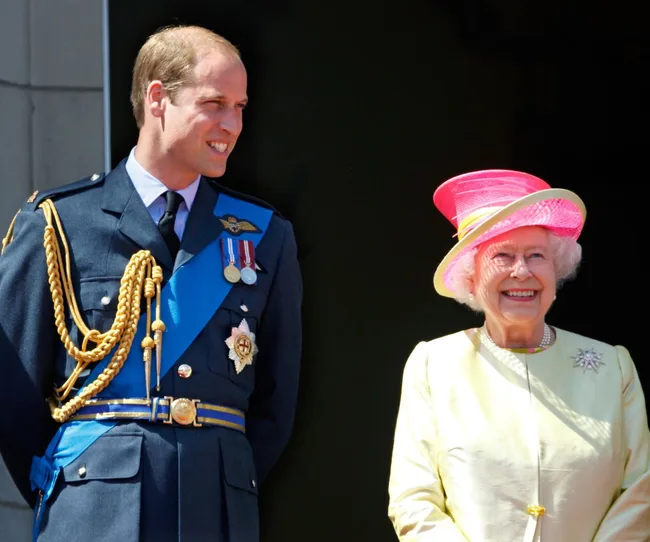 Prince in military attire with medals, standing beside a smiling elderly woman in a pink hat.