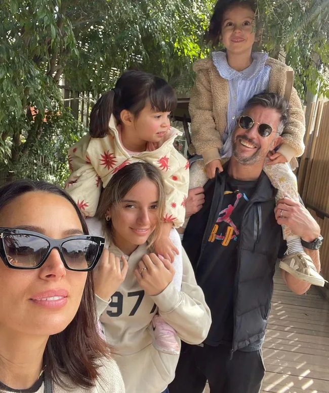 Family selfie with two adults and three children outdoors, one child on man's shoulders, others smiling, sunny day.