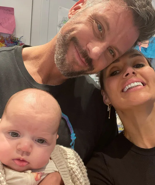 A man, woman, and baby close together, smiling for a selfie indoors.